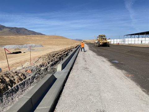 Freshly poured curb and gutter running alongside roadway subgrade, a motor grader working the lane.