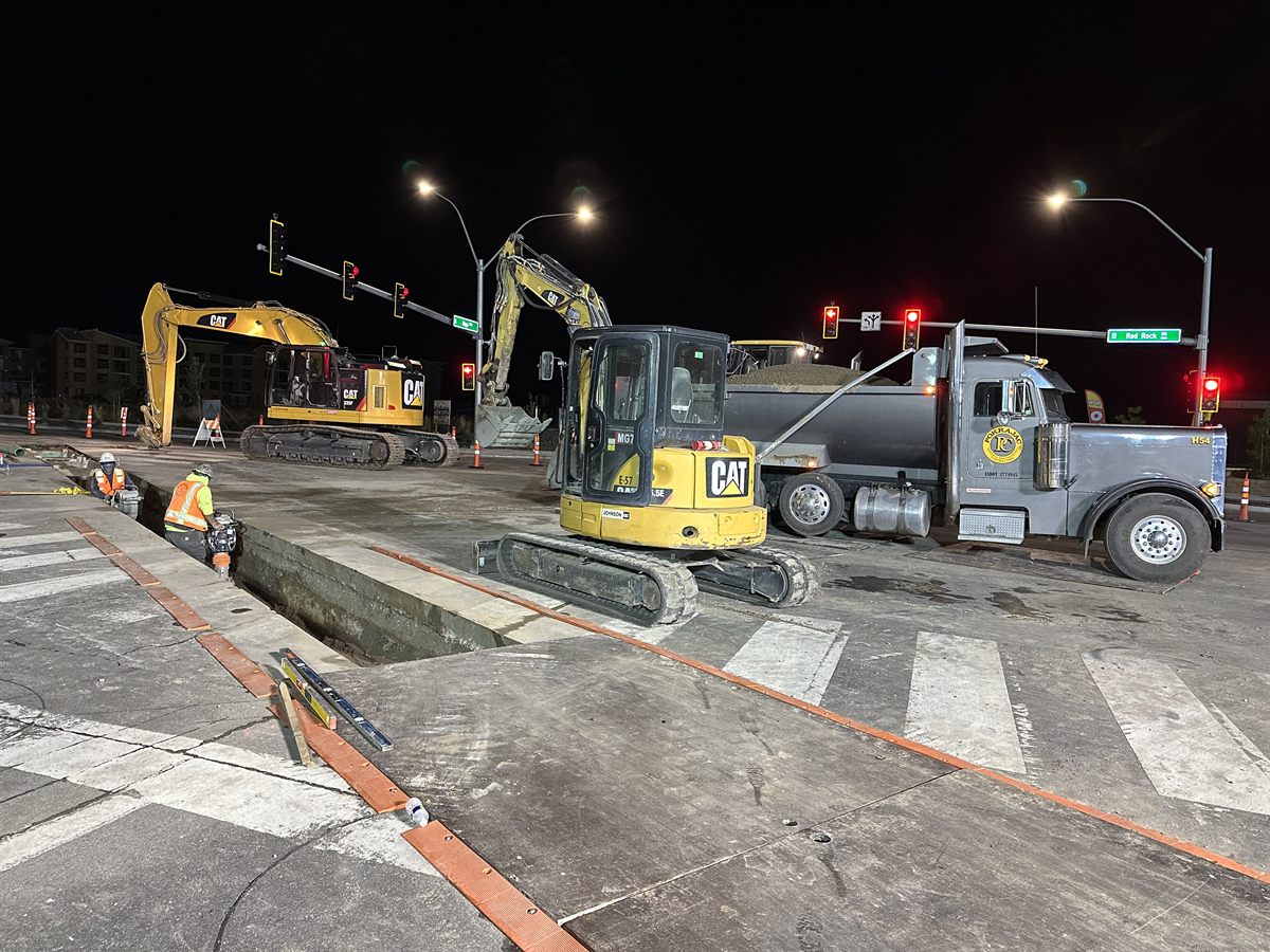 Night work at a live signalized intersection, two excavators and a Pokrajac truck under the street lights.
