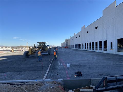 A motor grader working fine grade along a tilt-up distribution warehouse while crew set forms to line.
