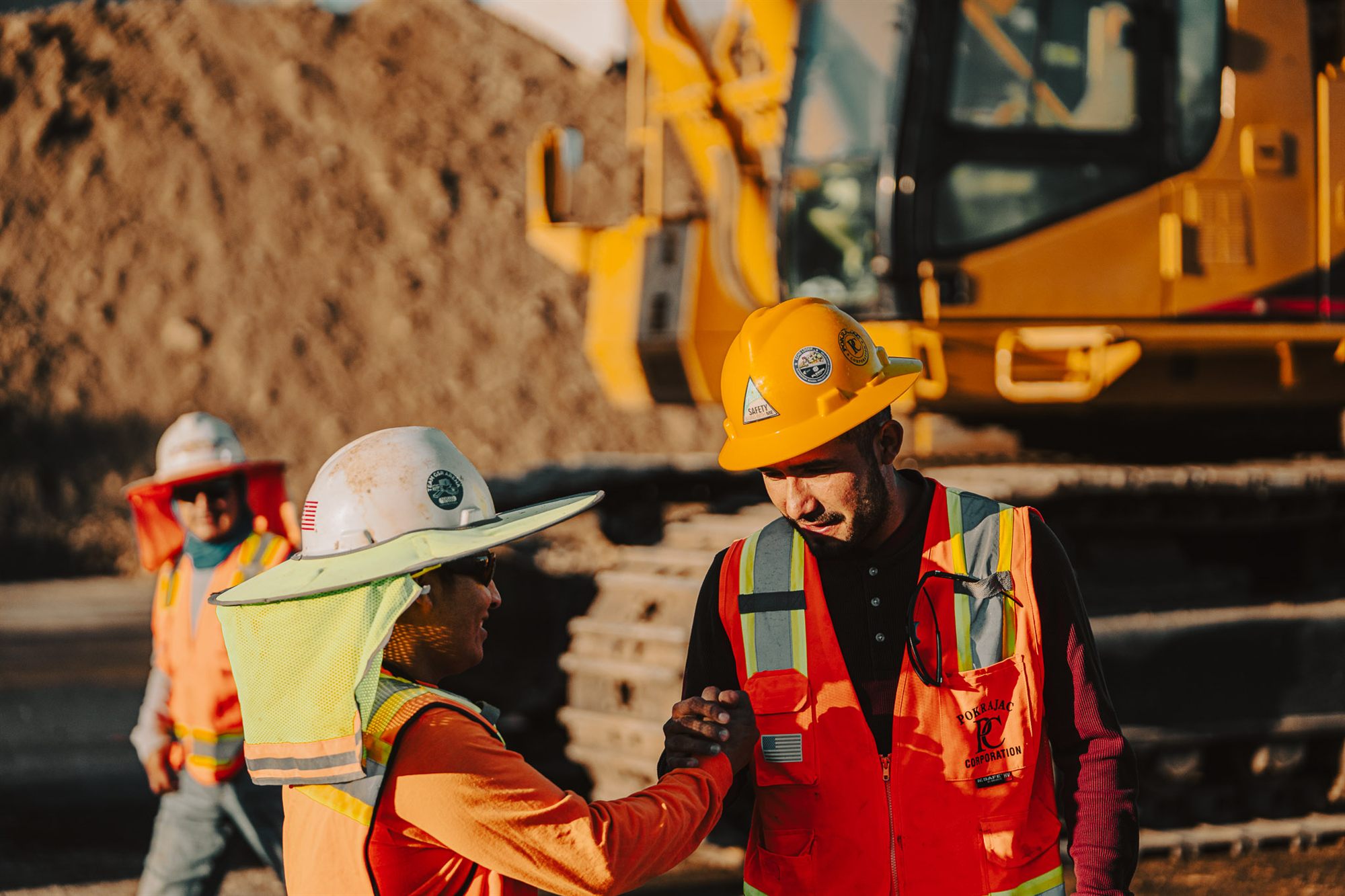 Two Pokrajac crew members greeting each other on site at golden hour, a Caterpillar excavator behind them.