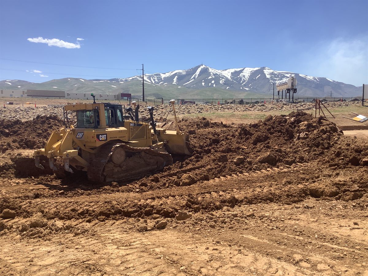A Caterpillar track dozer with rear rippers cutting grade, GPS mast up, snow-capped Sierra behind.